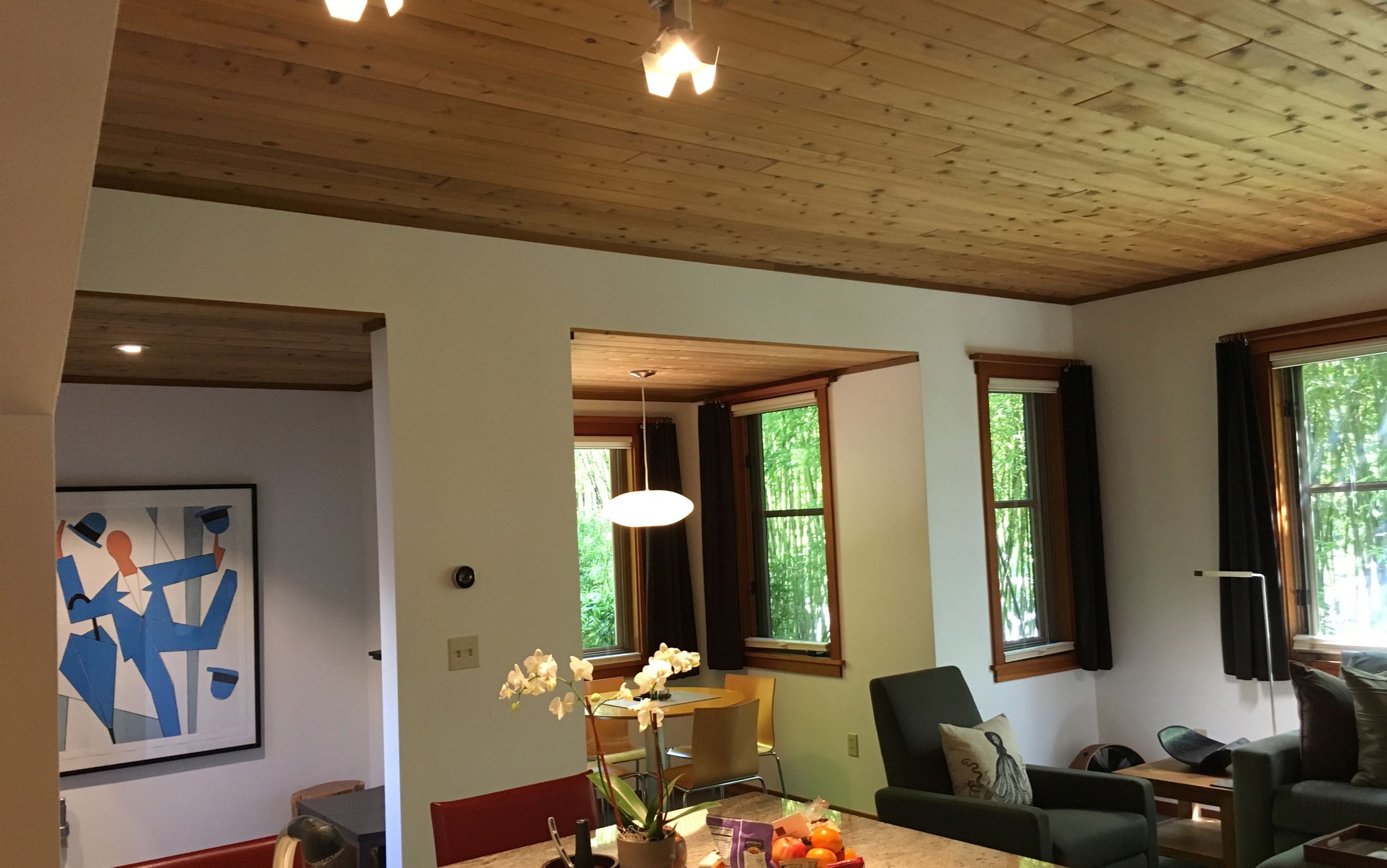 A spacious living room with wooden ceiling beams, large windows with curtains, a dining area with a table and chairs, and a painting on the wall.