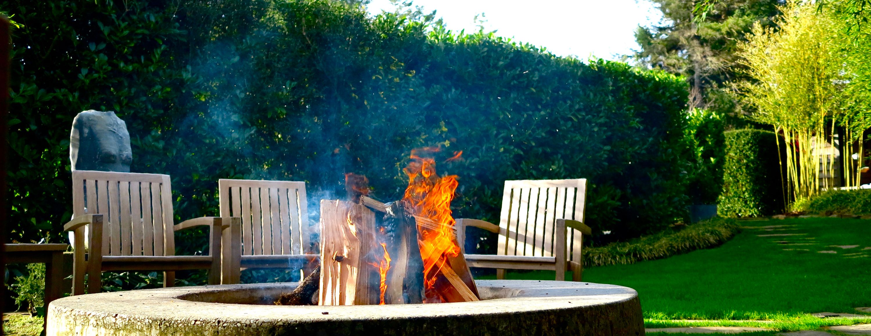 Een vuurplaats met brandende blokken hout, omringd door houten stoelen in een aangelegde tuin.