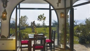 Ein Essbereich mit Tisch und Stühlen, Blick auf einen Garten und Bäume sowie ein Steinboden.
