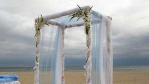 Ein Hochzeitbogen am Strand mit weißen und blauen Vorhängen und Blumendekorationen.