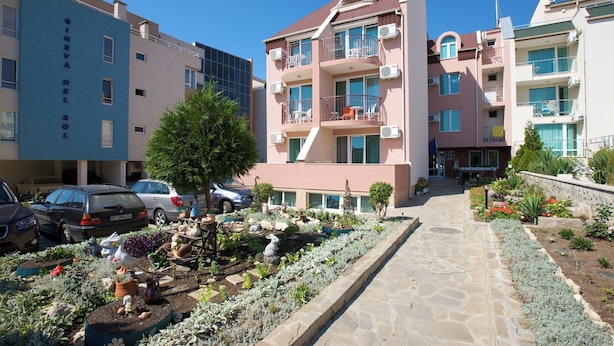 Ein mehrgeschossiges Wohngebäude mit Balkonen, einem Garten mit unterschiedlichen Pflanzen und Skulpturen sowie geparkten Autos.