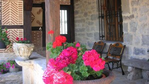 Een stenen gebouw met een houten veranda, een grote plantenbak met roze bloemen, en twee houten stoelen.