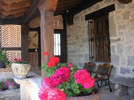 Un edificio de piedra con un porche de madera, una maceta grande con flores rosadas y dos sillas de madera.