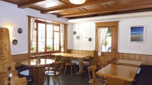 Ein Essbereich mit Holztischen und -stühlen, großen Fenstern mit Vorhängen und einer Holzbalkendecke.