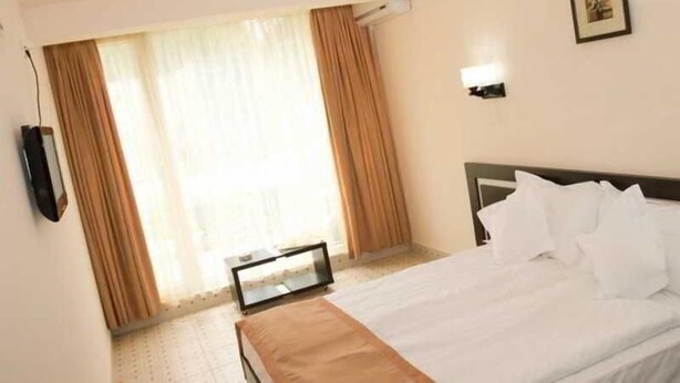 Una habitación de hotel con cama, cortinas, un televisor montado en la pared y un cuadro enmarco.