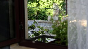 Vista a través de una ventana con cortina decorativa, un balcón con baranda de piedra y follaje en el exterior.