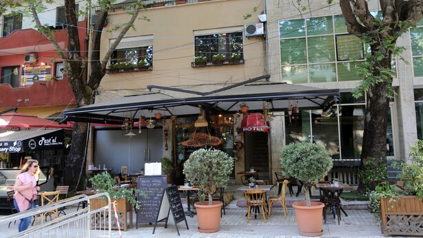 Un café al aire libre con mesas y sillas, un cartel y plantas en macetas.