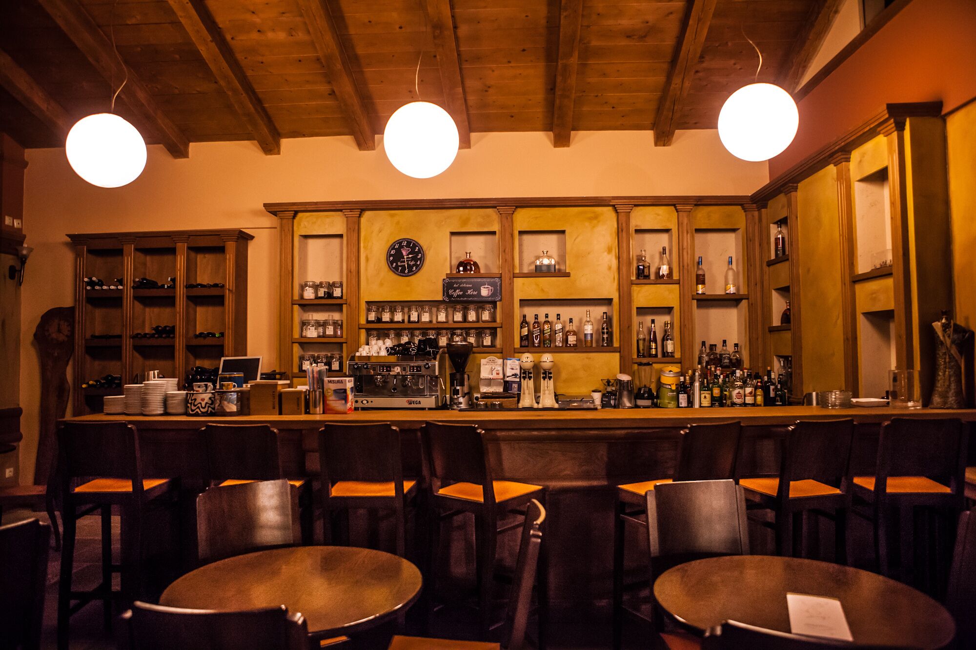 A bar with wooden shelves stocked with bottles, a coffee machine, and a clock on the wall. There are tables and chairs for seating.