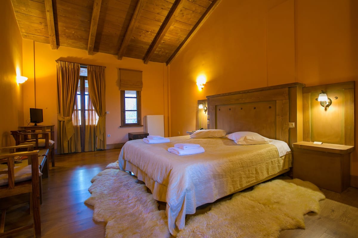 Ein Hotelzimmer mit Holzboden, einem Bett mit zwei Kissen, einem Nachttisch, einem Sessel, einem Schreibtisch mit Computer und einem Fenster mit Vorhängen.