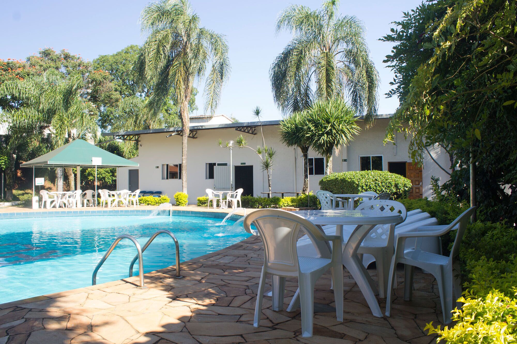 Uma área de piscina com cadeiras e mesas de plástico branco, uma área de estar coberta e um edifício com janelas e portas ao fundo.