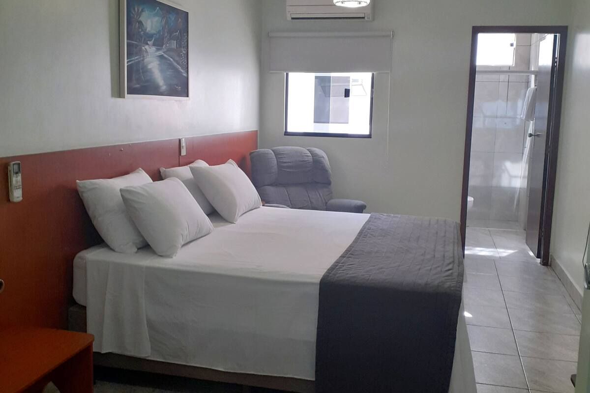 Ein Hotelzimmer mit Bett, Sessel, Schreibtisch, einem Bild an der Wand und einem Fenster.