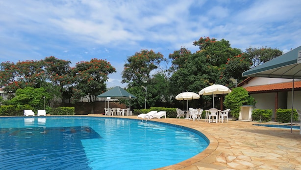 A swimming pool with stone tiles, surrounded by white lounge chairs and umbrellas, and trees with red flowers in the background. - Rio Claro Plaza Hotel (Rio Claro)