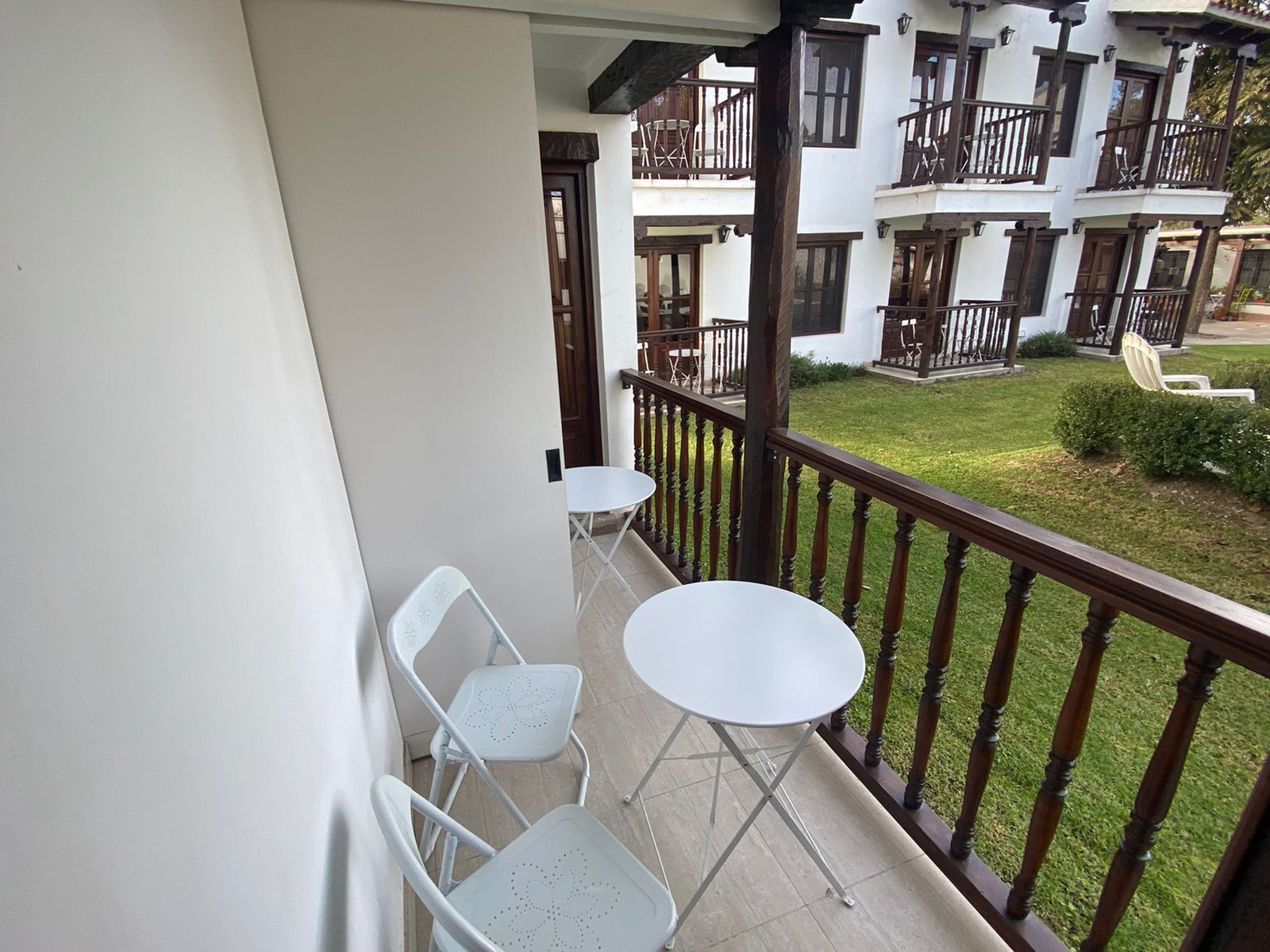 Un balcon avec deux tables et chaises blanches, une balustrade en bois et une vue sur un immeuble doté de balcons et d’une pelouse.