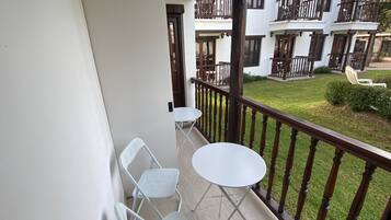 Un balcon avec deux tables et chaises blanches, une balustrade en bois et une vue sur un immeuble doté de balcons et d’une pelouse.