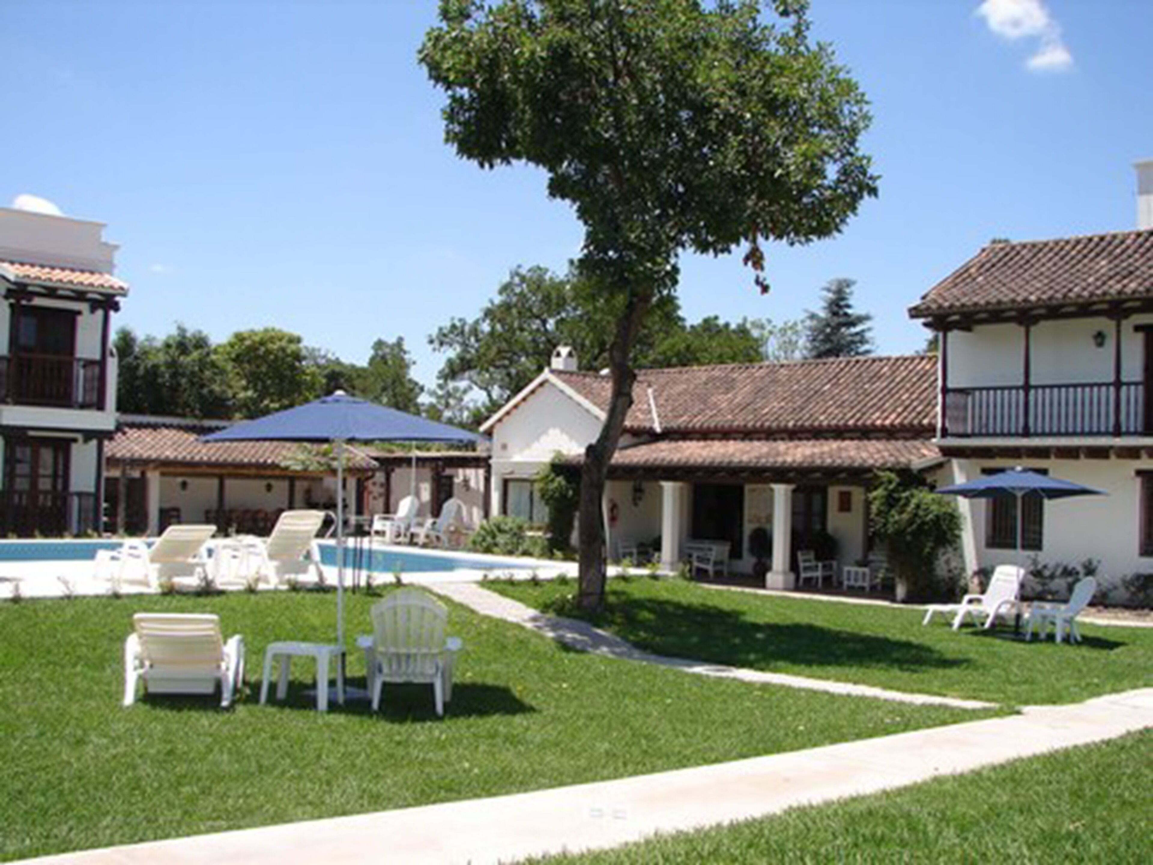 Un área de piscina con sillones y sombrillas, rodeada de edificios con techos de tejas y paredes blancas.