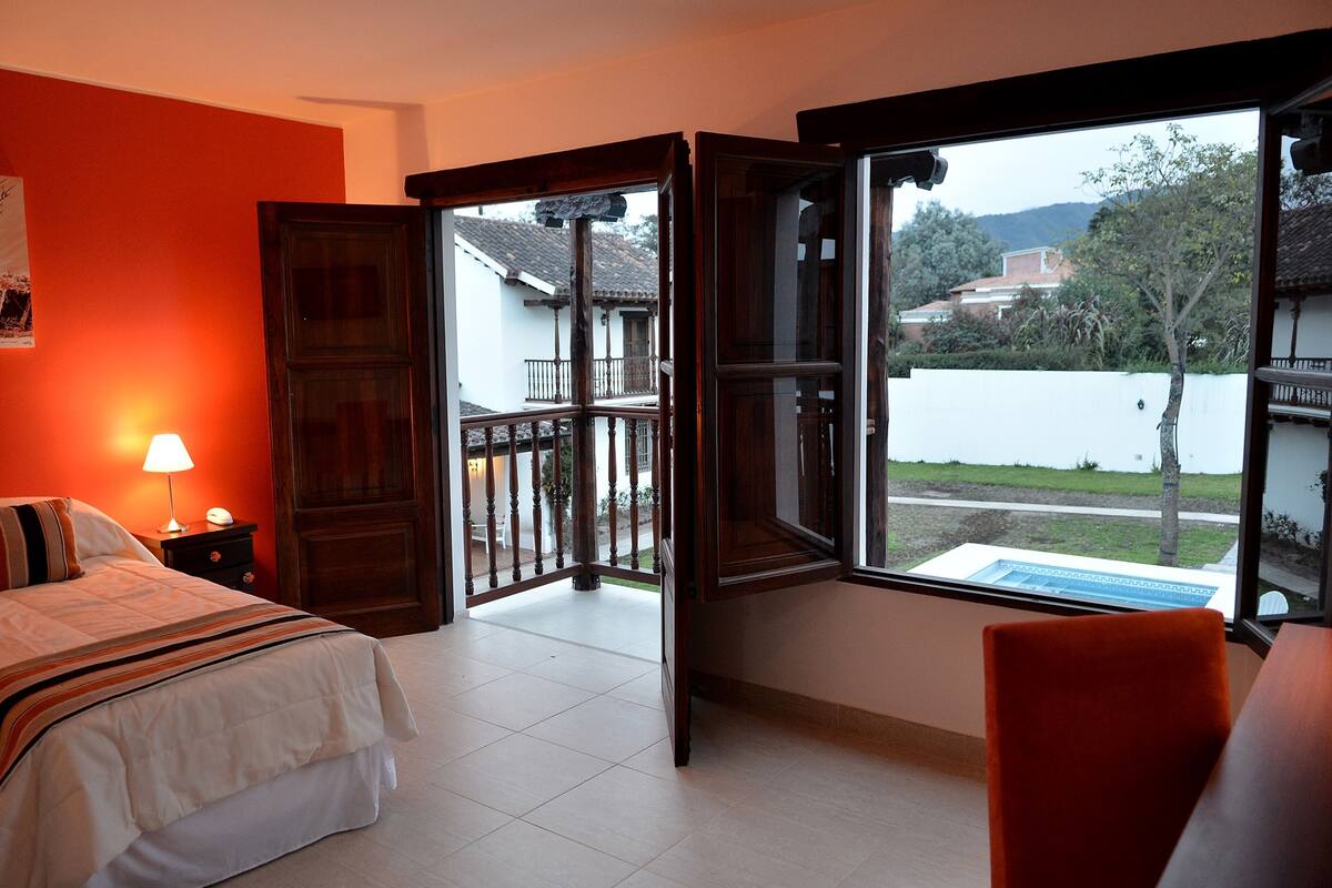 Ein Schlafzimmer mit einem Bett, einem Nachttisch, einer Lampe und einem Fenster mit Blick auf einen Balkon und Gebäude.