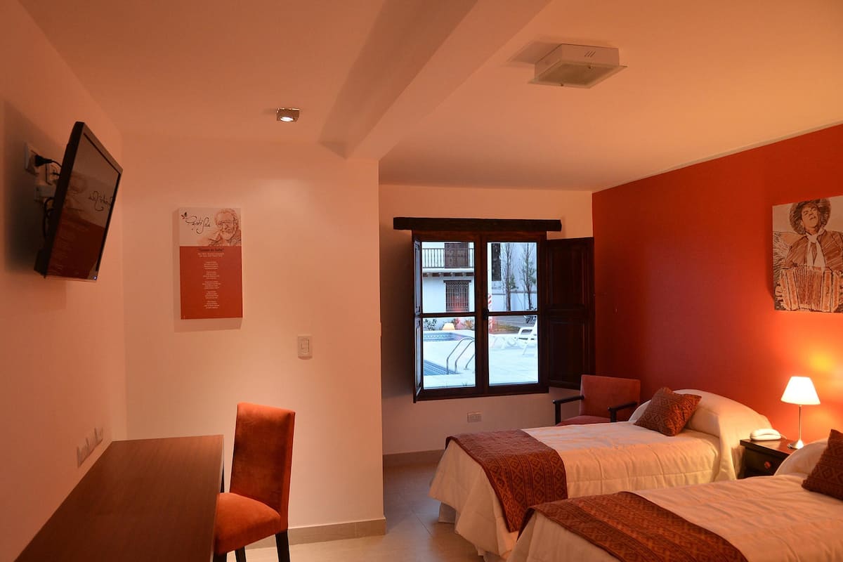 Ein Hotelzimmer mit zwei Betten, einem Schreibtisch, einem Stuhl, einem Fenster mit Blick nach draußen und einem Wand-Fernseher.