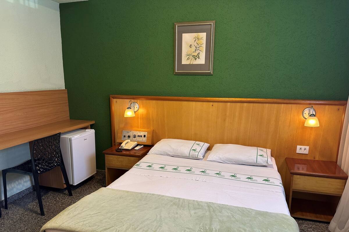 A hotel room with a bed, a desk, a refrigerator, a phone, and a nightstand with a lamp.