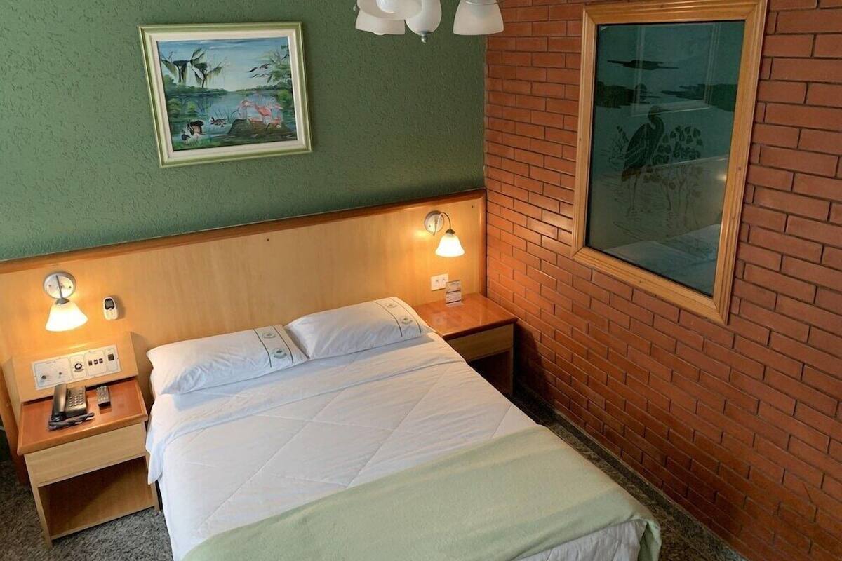 A hotel room with a bed, two bedside lamps, a nightstand with a phone, a framed picture on the wall, and a window with a view.