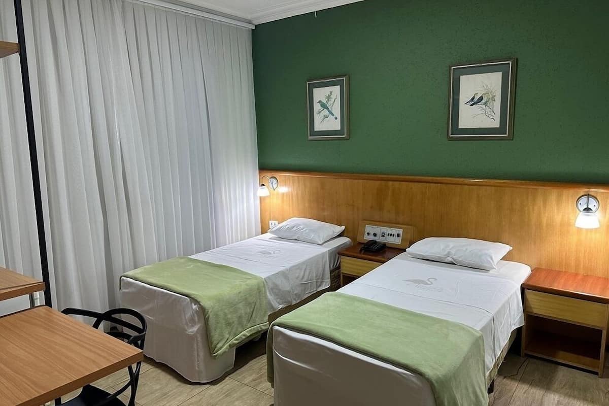 A hotel room with two single beds, a wooden headboard, a desk, a chair, and two wall-mounted lamps.