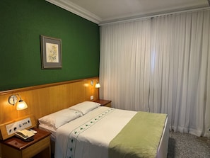 A hotel room with a bed, a nightstand with a telephone, a wall-mounted lamp, and a framed picture.