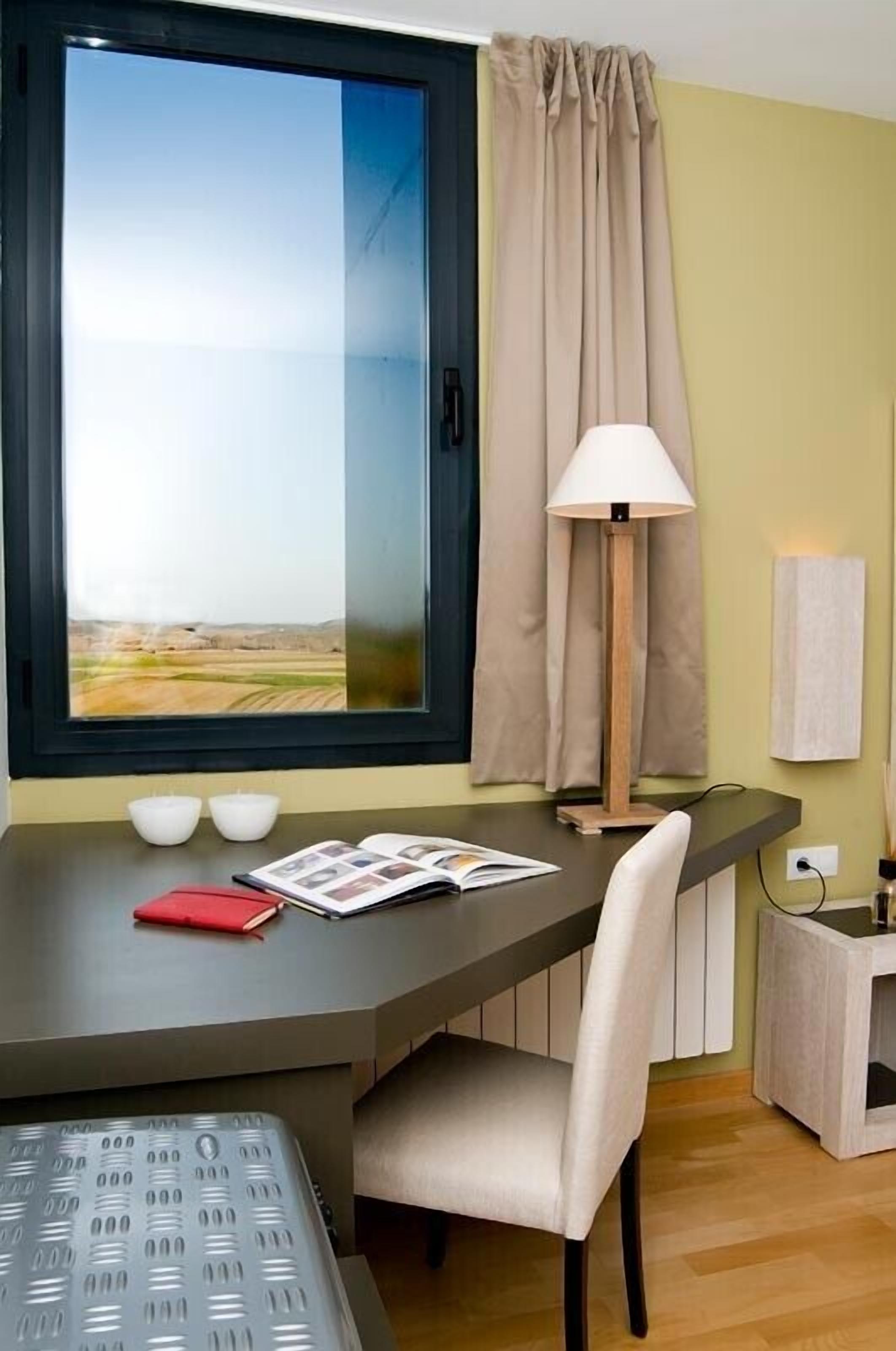 Una habitación con un ventanal grande, un escritorio con lámpara, una silla y una mesita de noche.