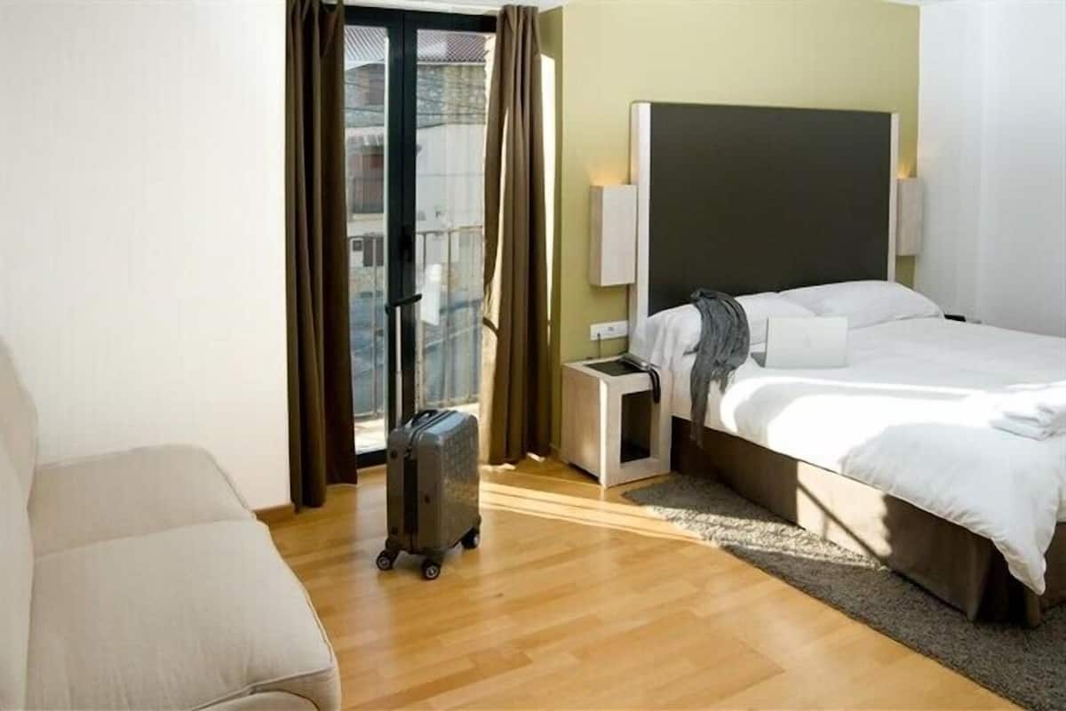 A hotel room with a bed, a small bedside table, a suitcase, and a view of the outside through a sliding door.