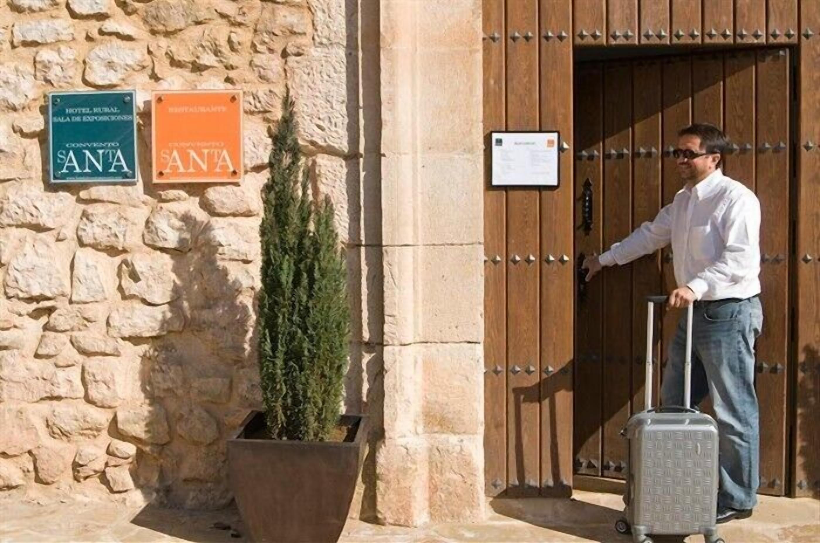 Un hombre con maleta en la entrada de un edificio de paredes de piedra y puerta de madera. Se observan letreros que indican un hotel y un restaurante llamado 'SANTA'.
