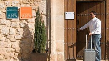 Un hombre con maleta en la entrada de un edificio de paredes de piedra y puerta de madera. Se observan letreros que indican un hotel y un restaurante llamado 'SANTA'.