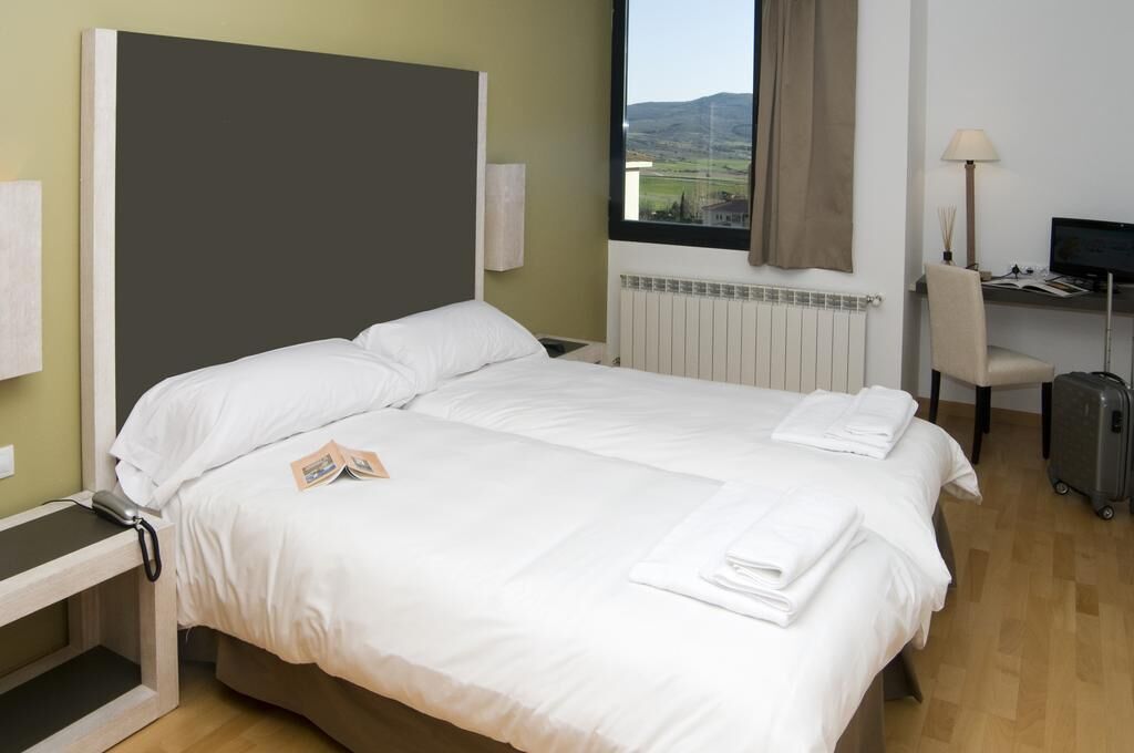 A hotel room with a bed, a desk, a chair, a lamp, and a view of the countryside.