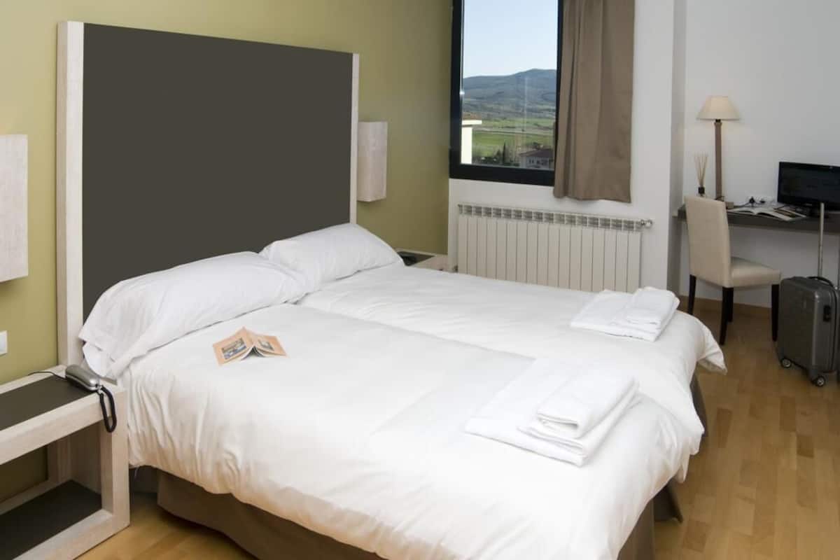 A hotel room with a bed, a desk, a chair, a lamp, and a view of the countryside.