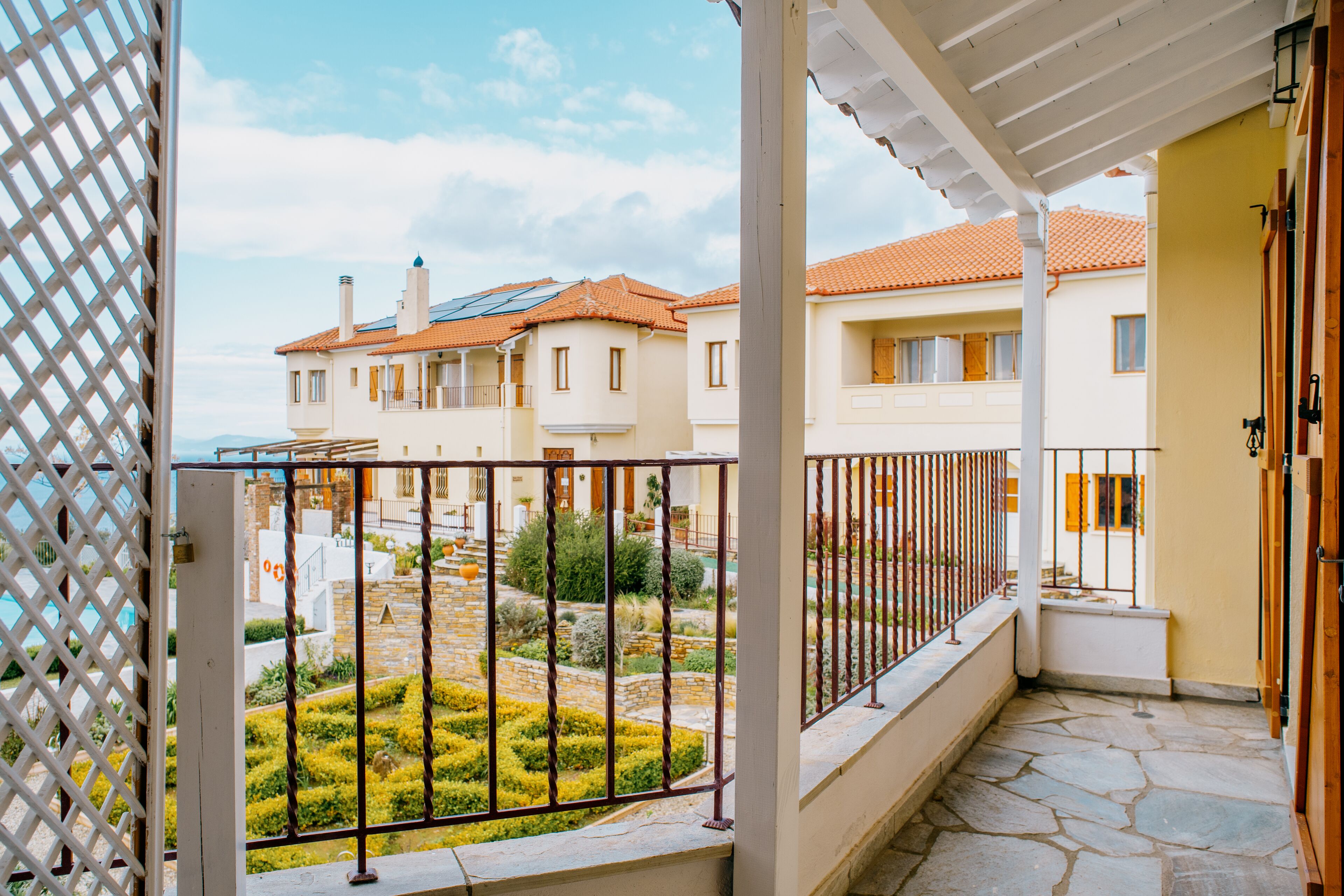 A balcony with a stone floor, a white railing, and a view of a courtyard with neatly trimmed bushes and a small tree.