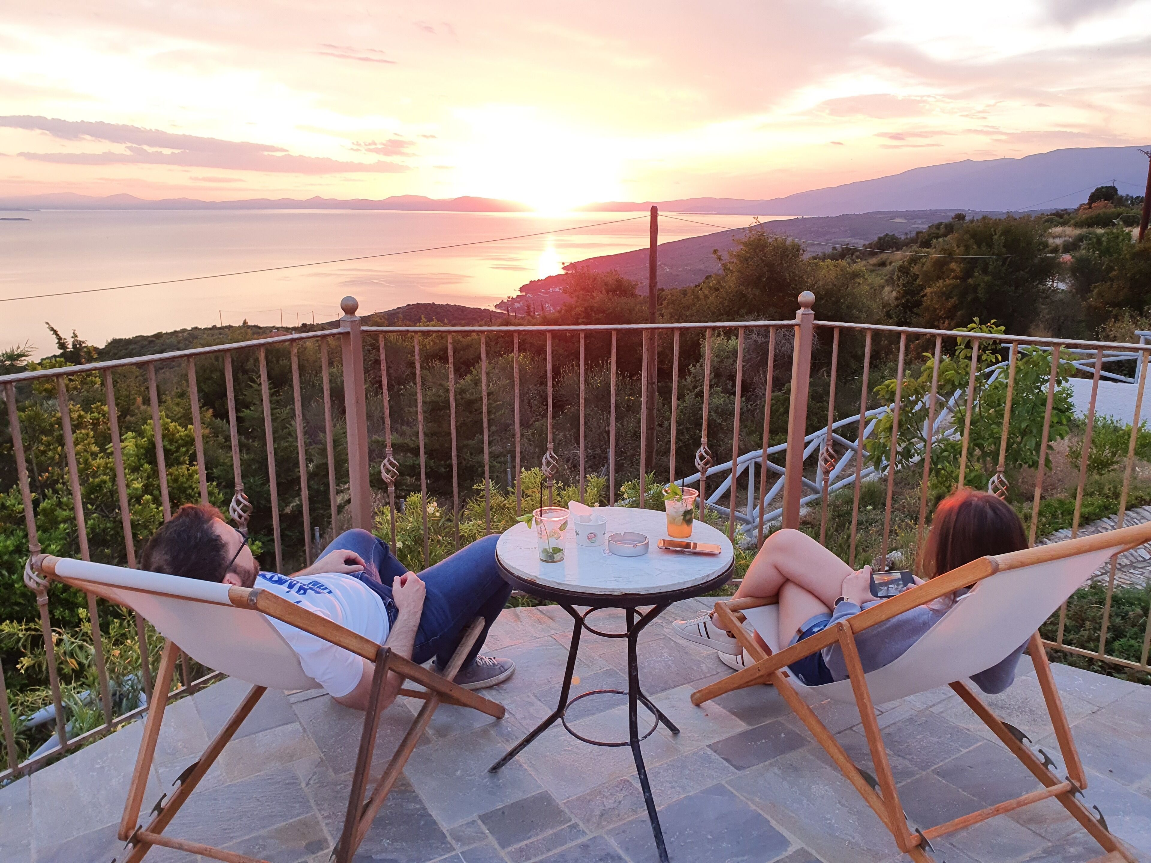 Dos personas recostadas en sillas al aire libre, junto a una mesa con bebidas, contemplando una vista panorámica del mar y las colinas al atardecer.