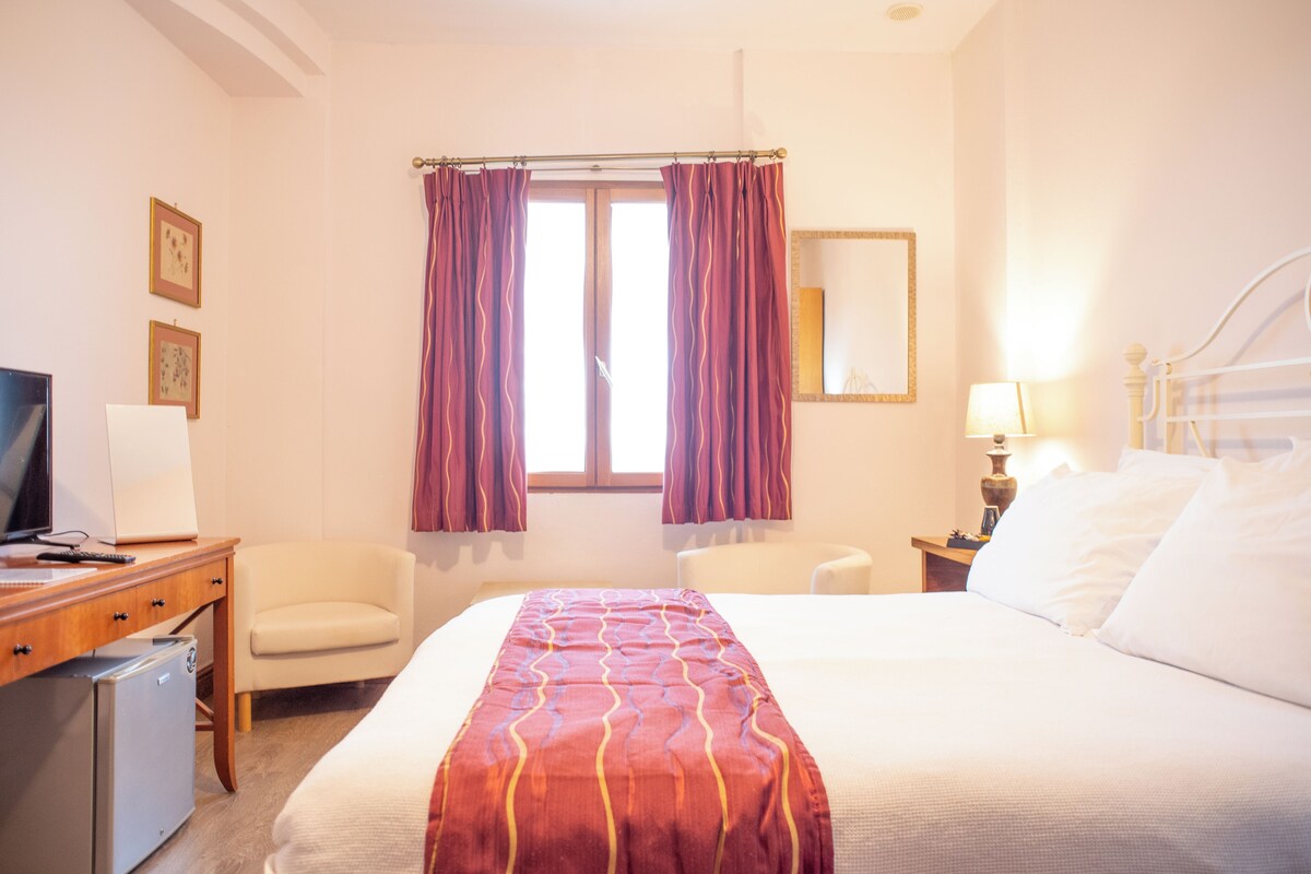 Habitación de hotel con cama, escritorio con computadora, silla, televisor y ventana con cortinas.