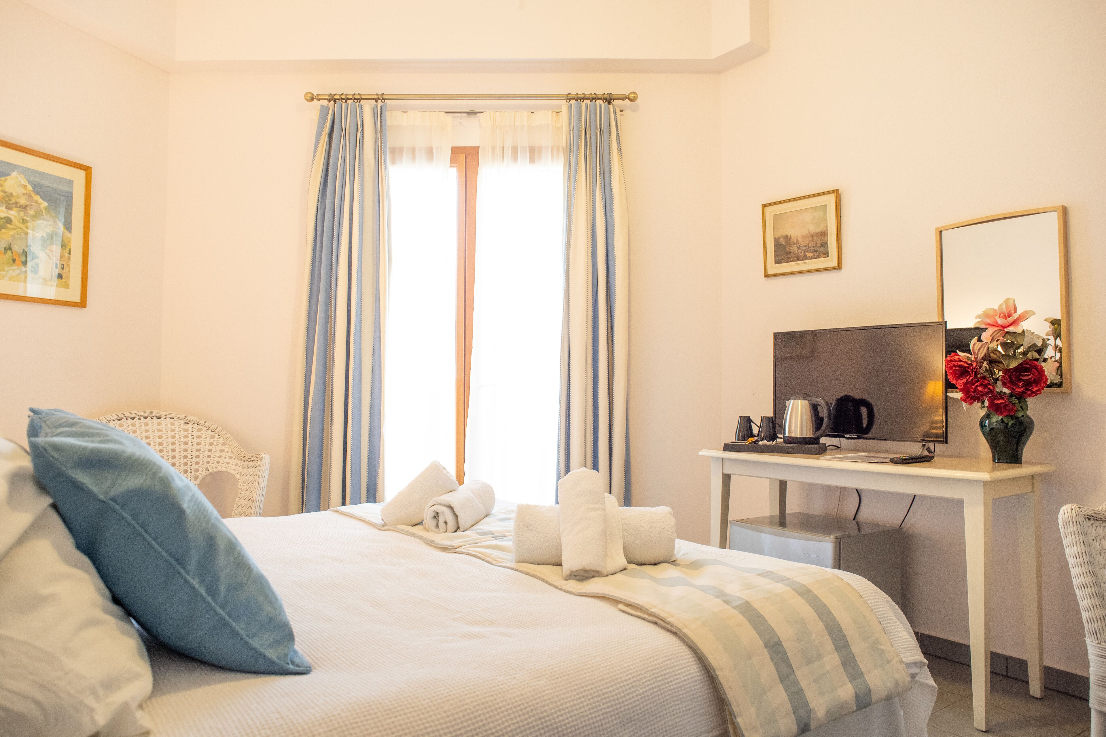 A hotel room with a bed, a desk with a television, a chair, and a vase with flowers.