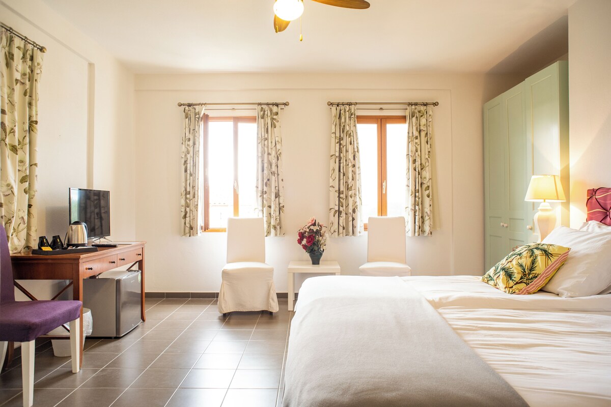 Habitación de hotel con una cama, un escritorio con computadora, dos sillas, una mesita con un florero y una lámpara.