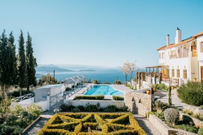 Zona de piscina con vista al mar, rodeada de vegetación y un edificio blanco con balcones.
