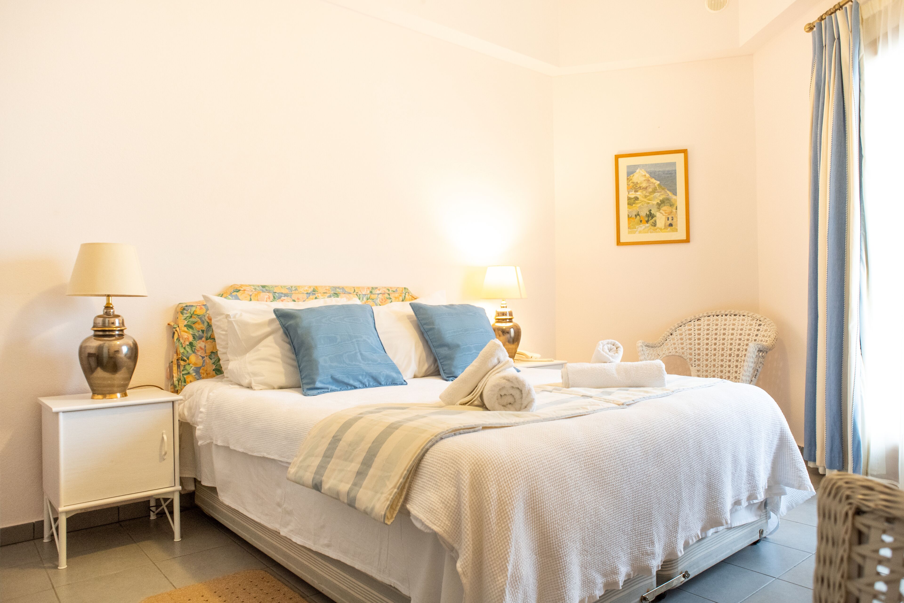 A neatly made bed with white bedding and blue pillows, a small bedside table with a lamp, a wicker chair, and a framed picture on the wall.