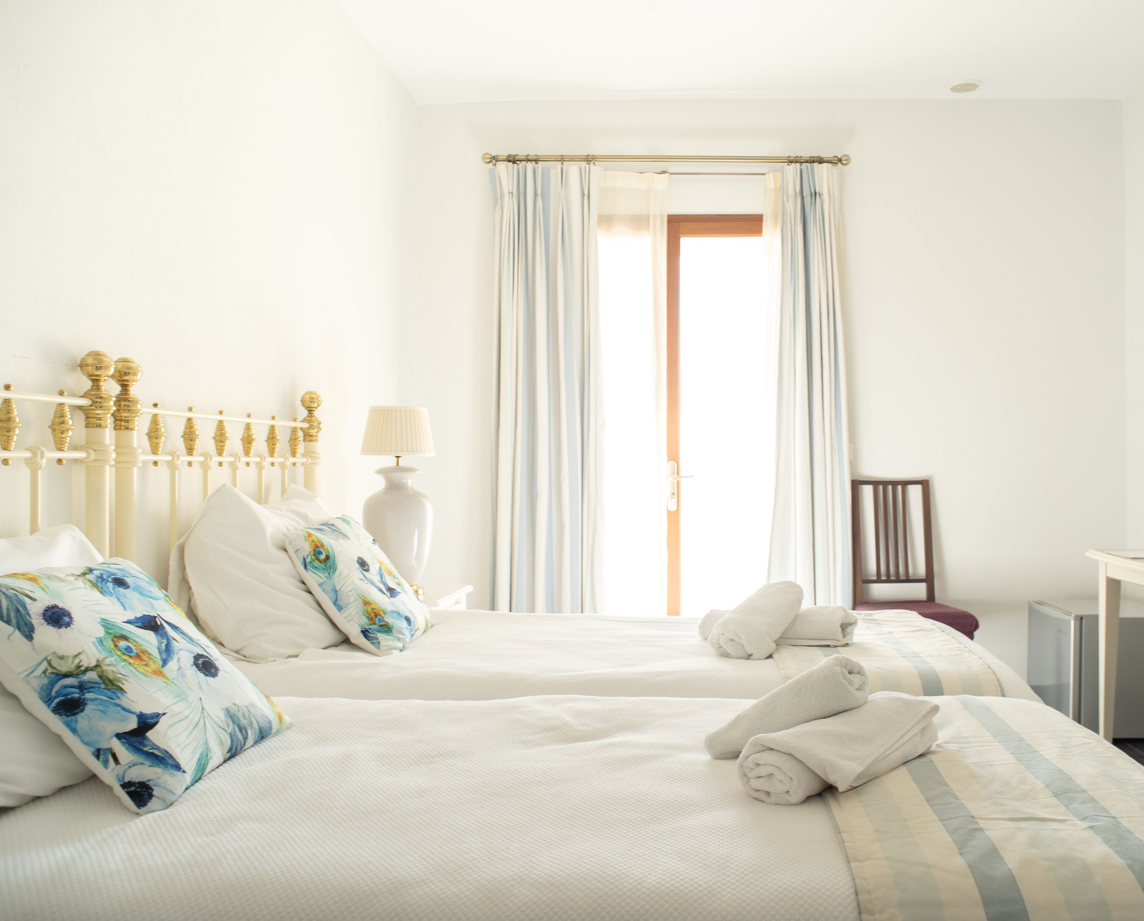 Habitación de hotel con dos camas, una mesita de noche con lámpara y vistas al exterior a través de una ventana con cortinas.