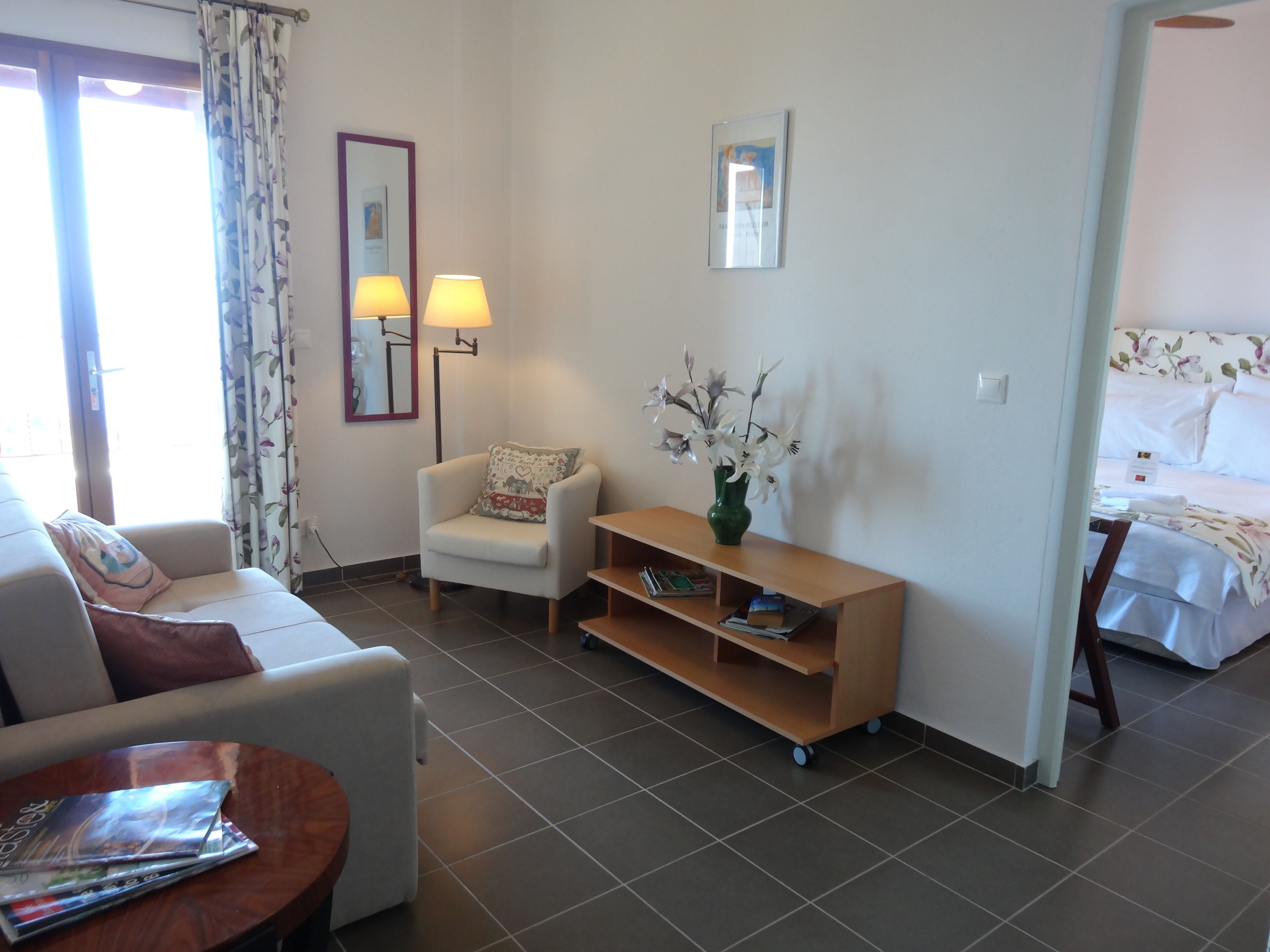 A modern hotel room with a sofa, armchair, wooden side table, and a bed with a floral bedspread.