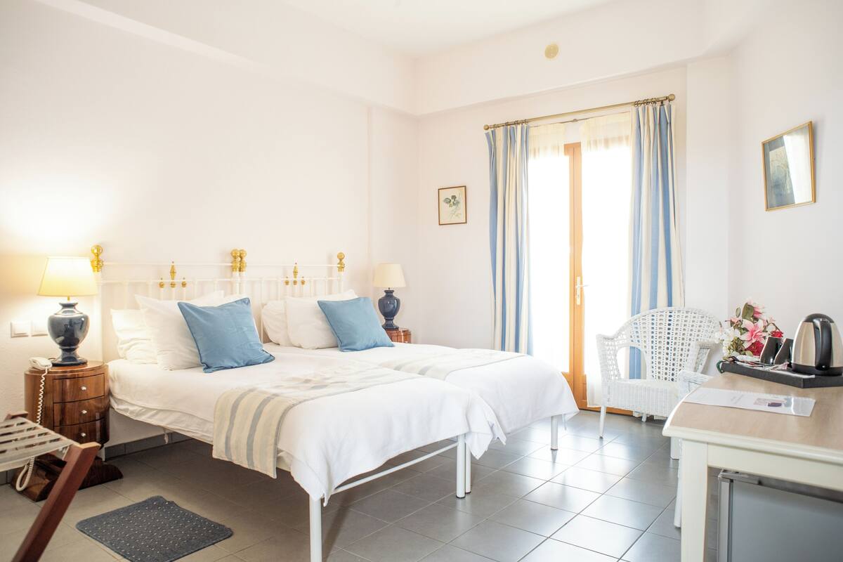 A hotel room with a double bed, two bedside tables with lamps, a chair, a desk with a kettle, and a window with curtains.