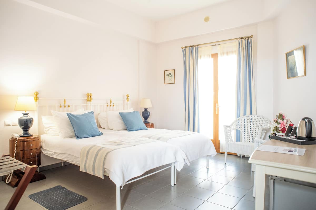 A hotel room with a double bed, two bedside tables with lamps, a chair, a desk with a kettle, and a window with curtains.