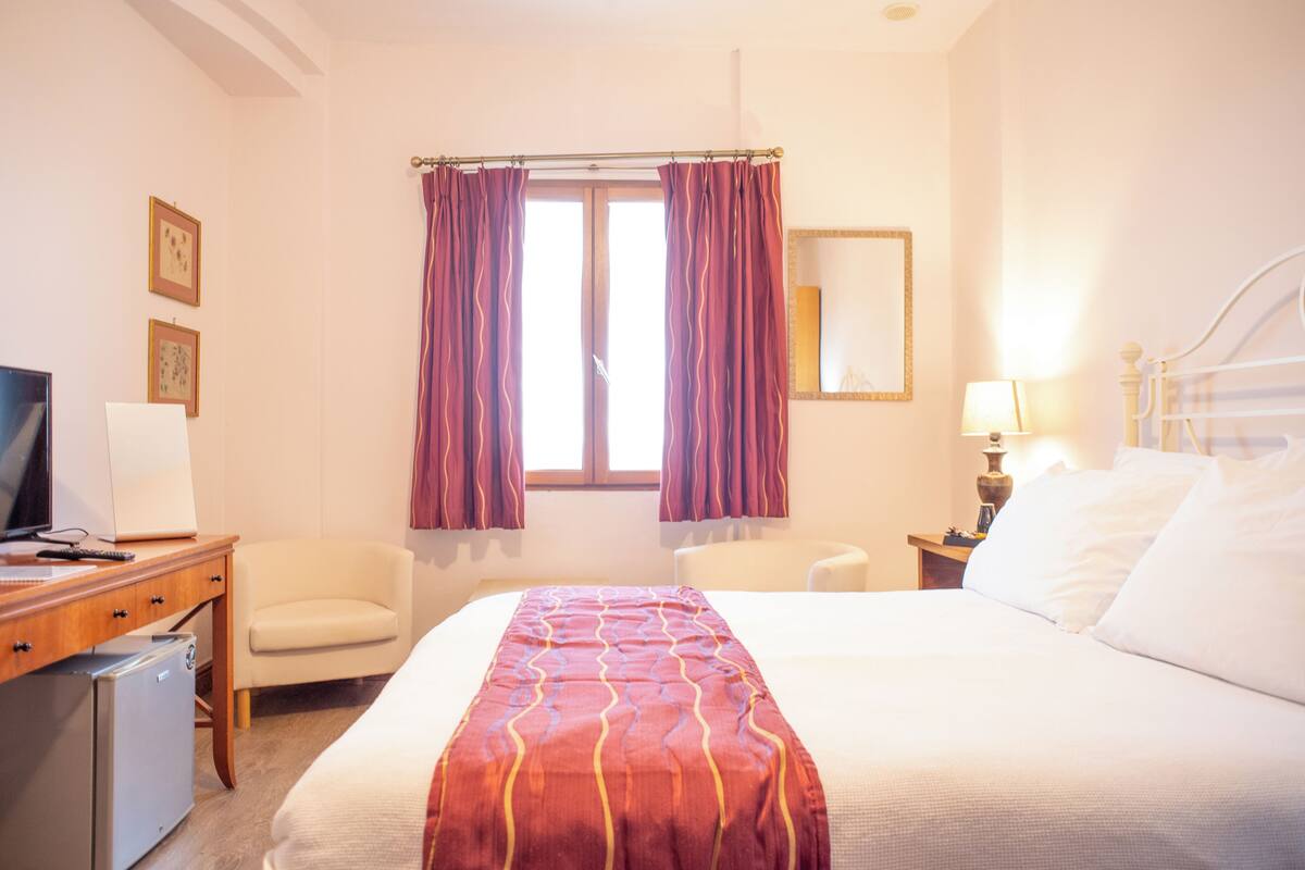 A hotel room with a bed, a desk with a computer, a chair, a TV, and a window with curtains.