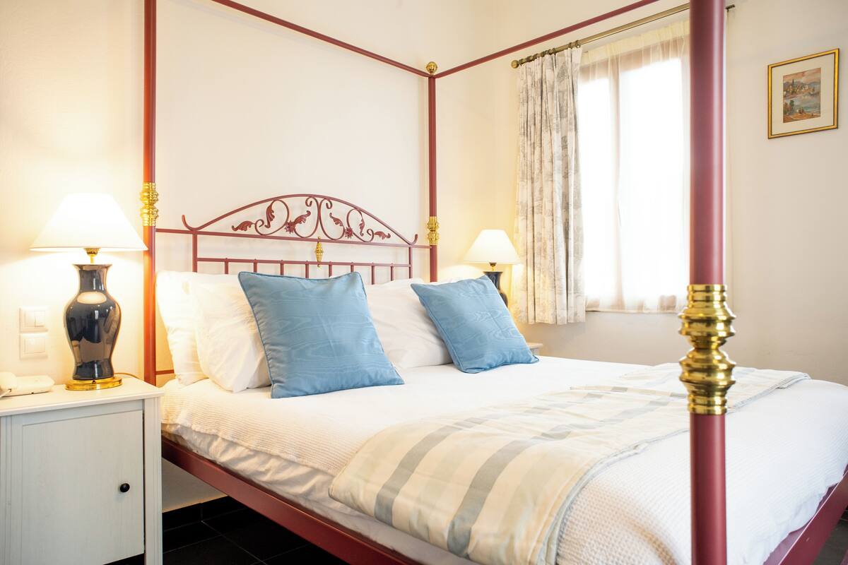 A four-poster bed with a white headboard and blue pillows, a nightstand with a lamp, and a window with curtains.