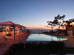 Zona junto a la piscina con un pabellón, sillas de descanso y vista al mar al anochecer.