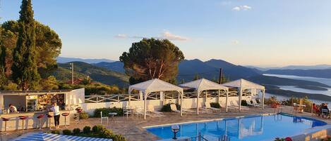 Un área de piscina con vistas a montañas y un lago, rodeada de mobiliario exterior y un bar.