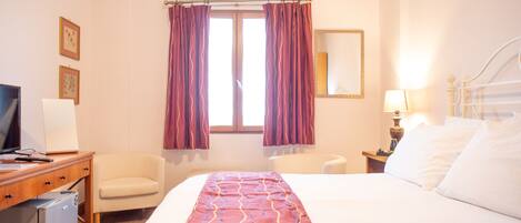 A hotel room with a bed, a desk with a computer, a chair, a TV, and a window with curtains.