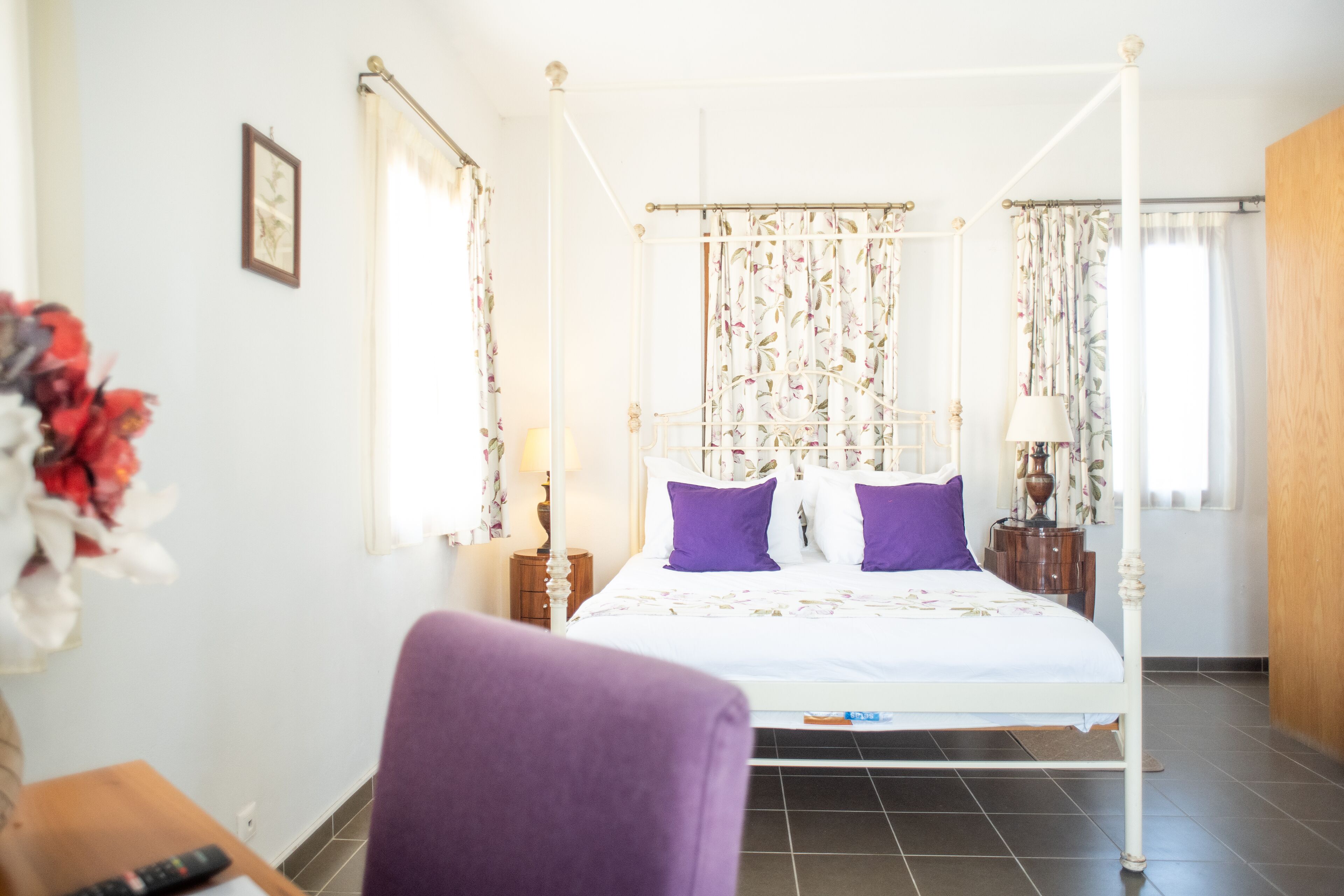 A four-poster bed with white bedding and purple pillows, a wooden nightstand with a lamp, and a chair in a room with tiled flooring and floral curtains.