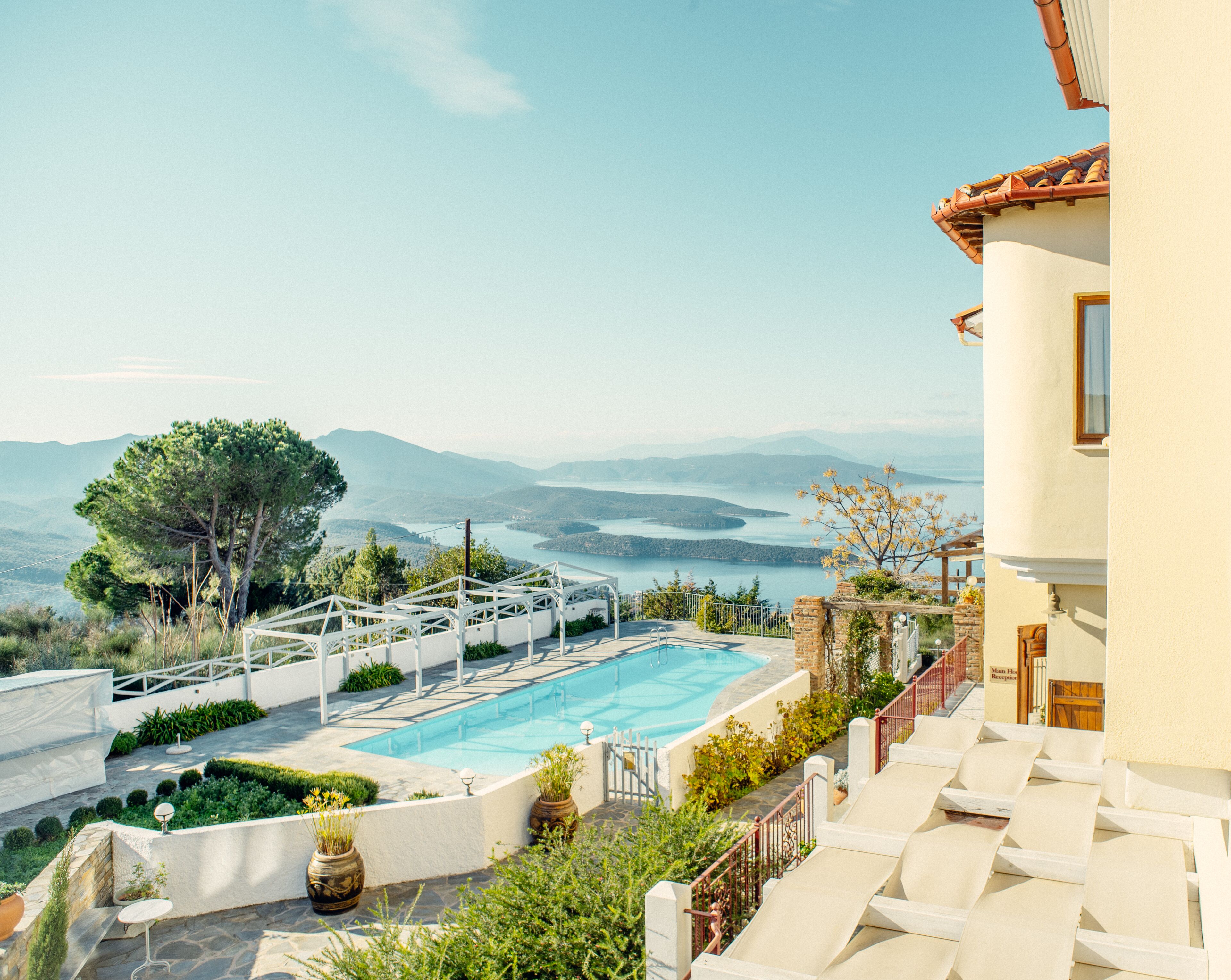 A pool area with a view of a lake and mountains, surrounded by greenery and a white building.