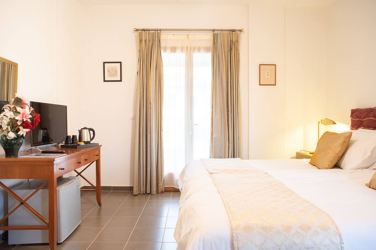 A hotel room with a bed, a desk with a computer, a small refrigerator, and a window with curtains.